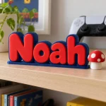 Prénom 3D Mario personnalisé à poser : lettres rouges et bleues sur une étagère d'enfant avec une manette en arrière-plan